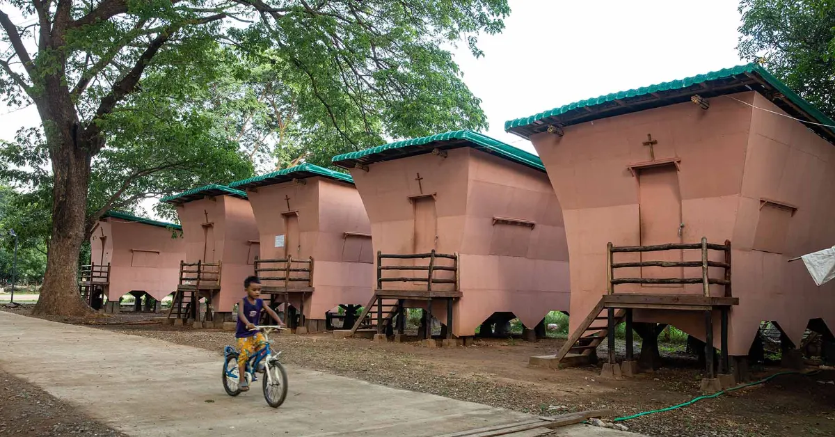 A child rides a bicycle past a row of five pink structures with teal roofs and crosses mounted on their walls, elevated on wooden platforms beneath a large tree.