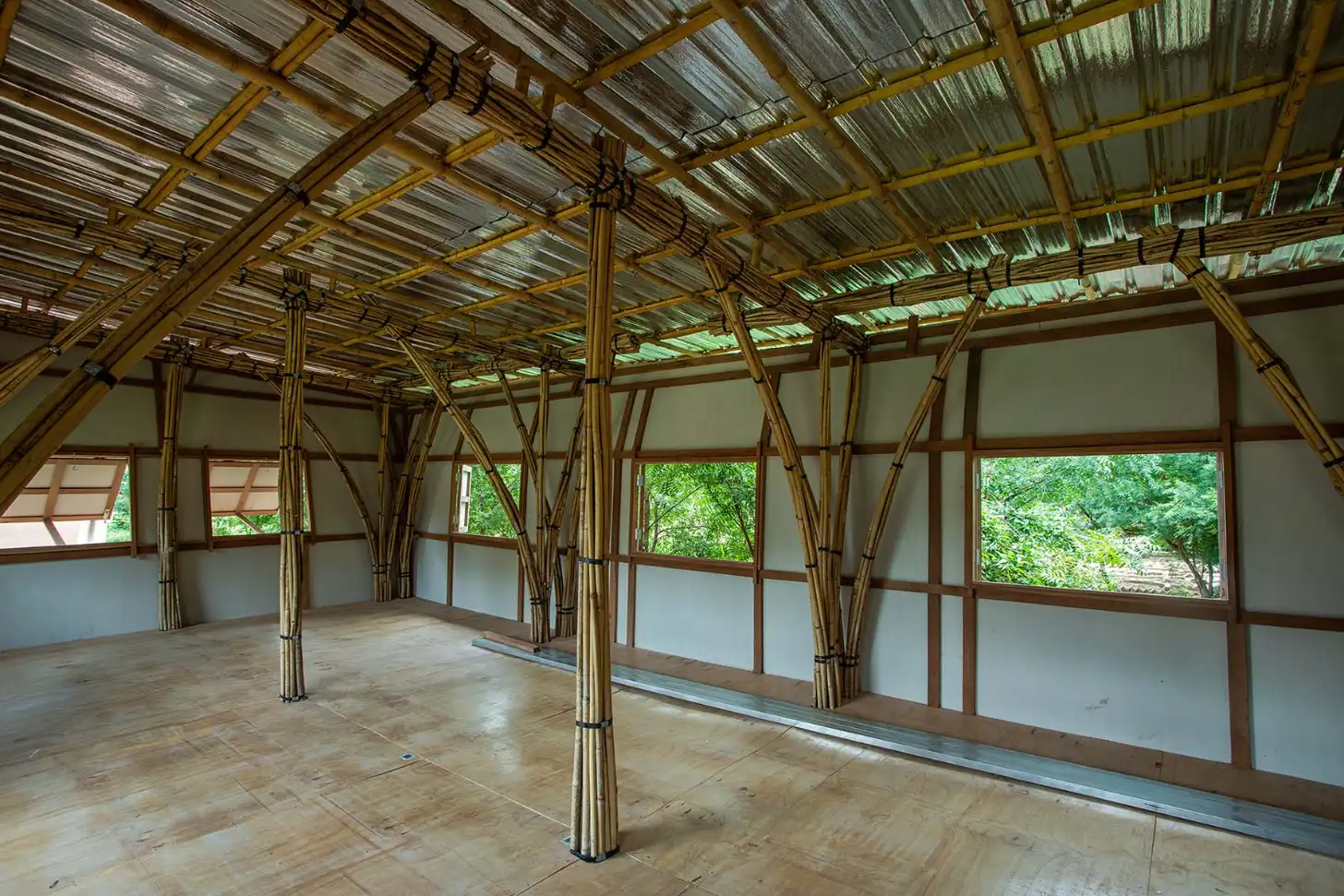 Interior of a large open structure with bamboo support columns, corrugated metal roof, and windows along the walls overlooking green foliage outside.