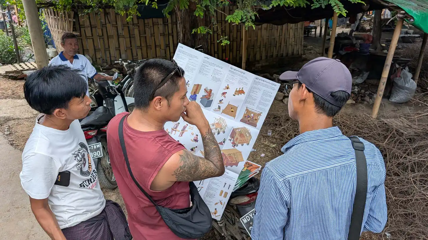 Three men examine a large printed document with illustrations and text in an outdoor courtyard setting. Motorcycles and wooden fencing are visible in the background.