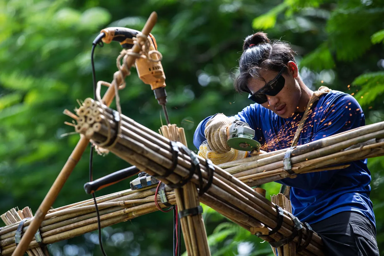 A worker wearing dark sunglasses and a blue shirt grinds metal on bamboo scaffolding, with sparks flying from the tool. A yellow pneumatic tool hangs from above against a blurred green foliage background.