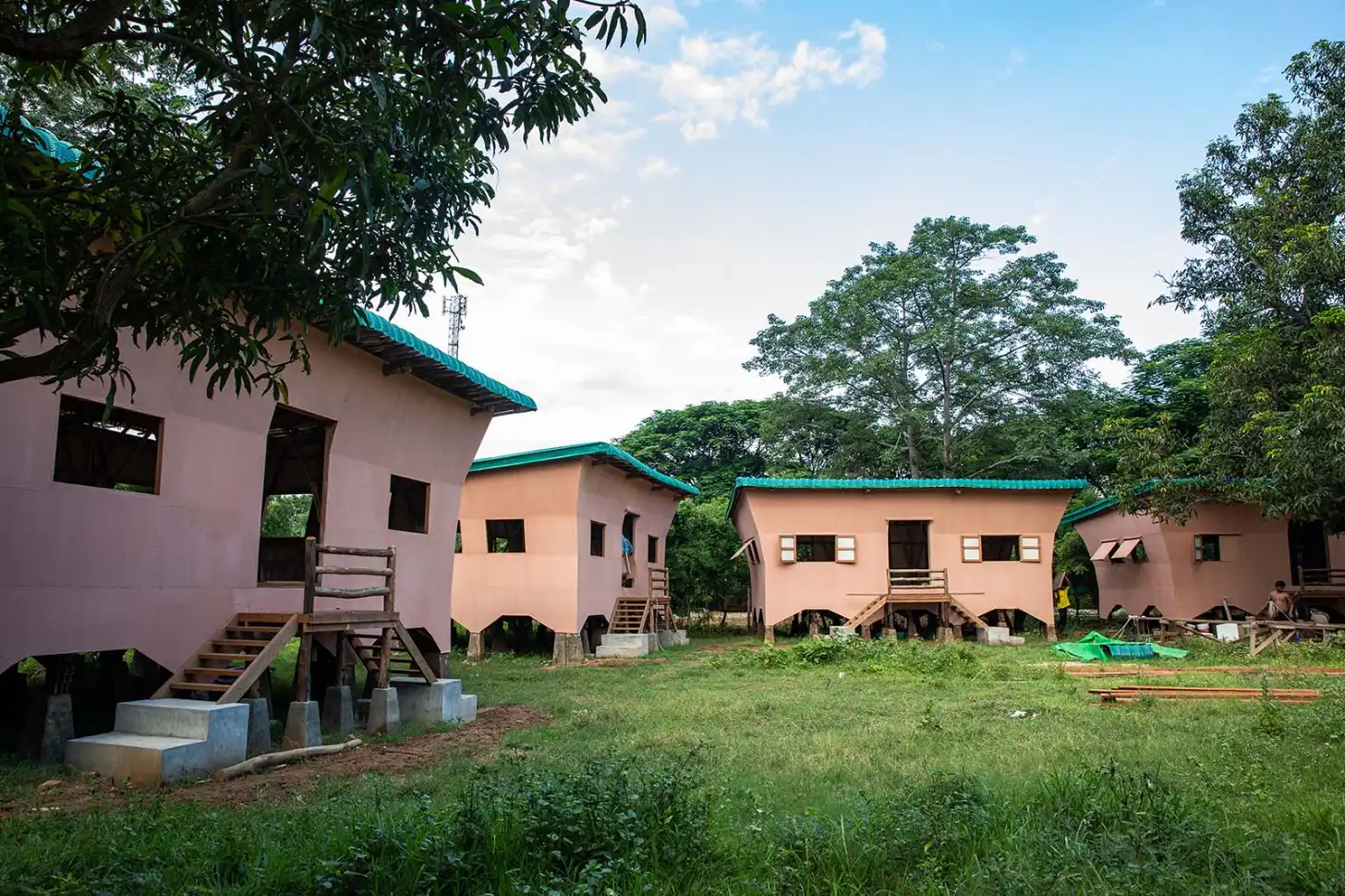 Several single-story houses with pink and peach-colored walls and turquoise metal roofs are elevated on concrete pillars in a grassy clearing surrounded by tall trees.