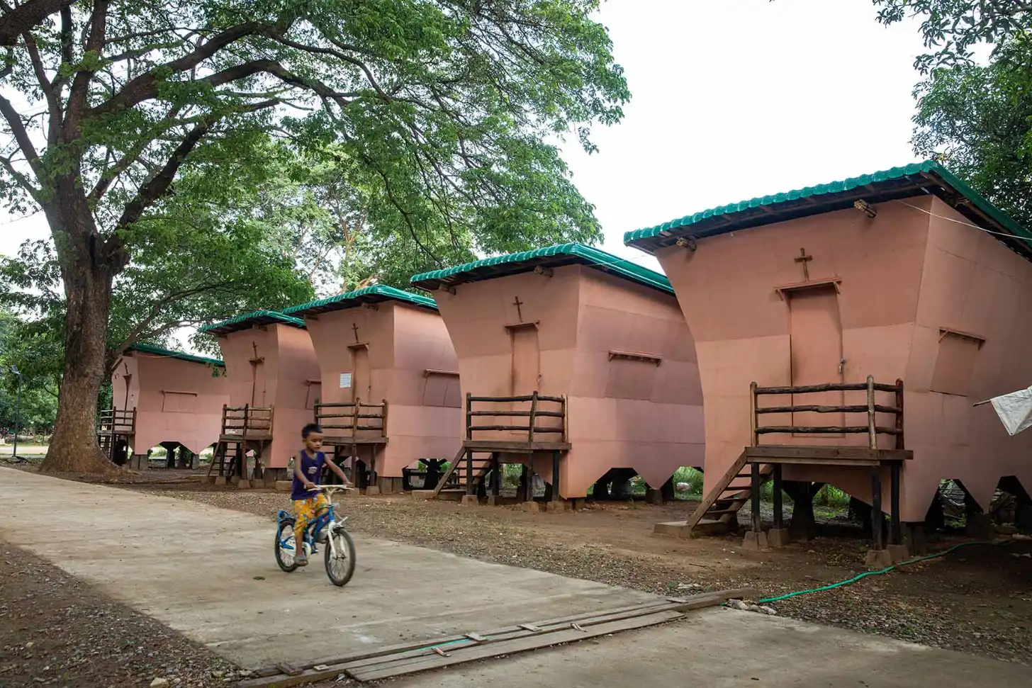 A child rides a bicycle past a row of pink buildings with green roofs and crosses on their facades, standing on stilts beneath a large tree.