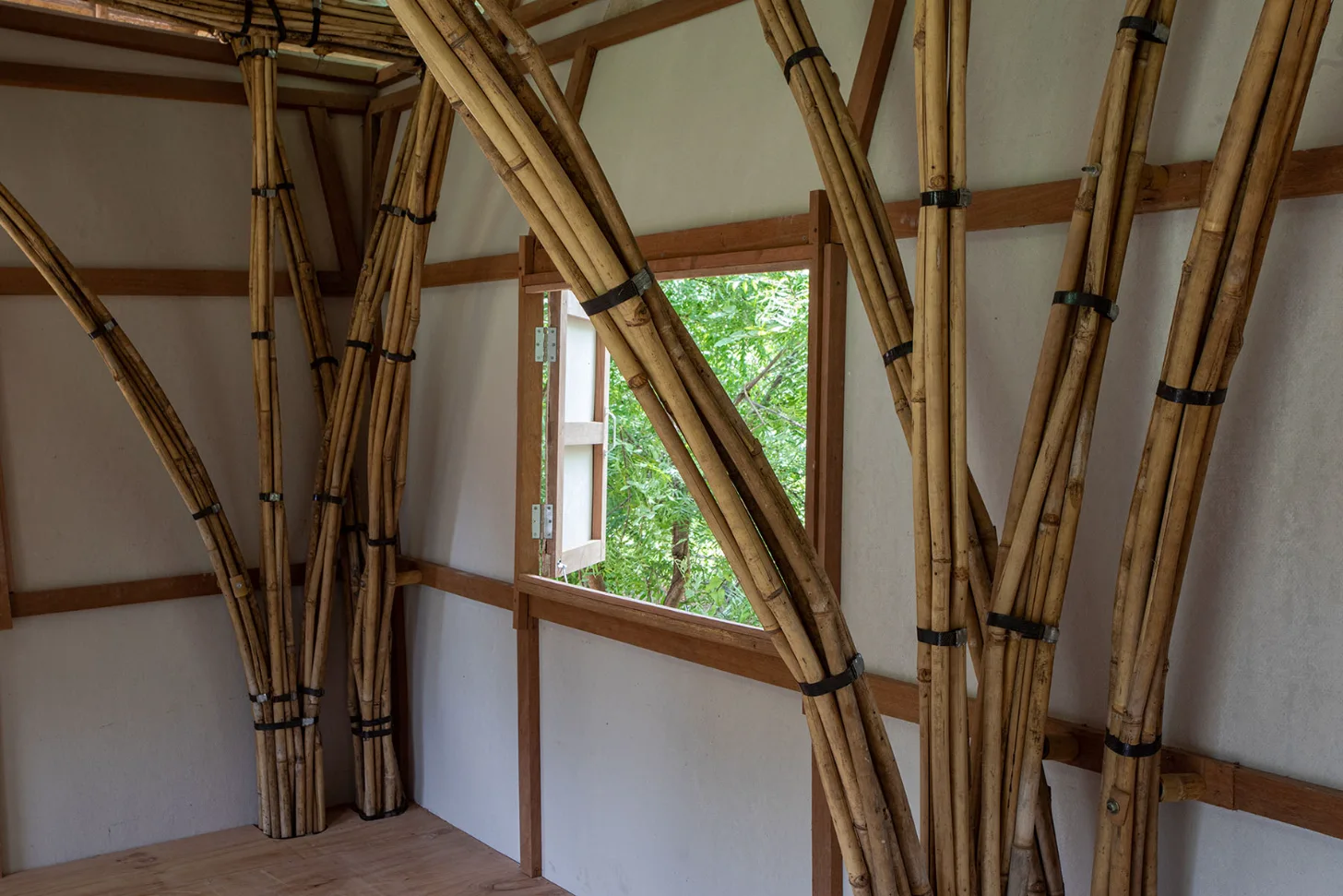 Interior room with bundled bamboo poles tied with black straps forming an architectural structure, white walls, wooden beams, and a window framing green foliage outside.