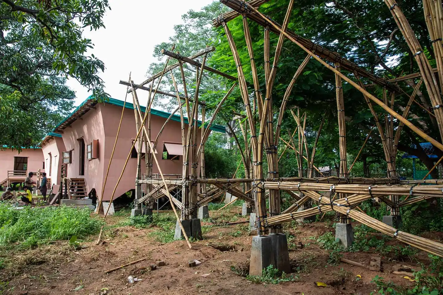 A pink building with a teal roof stands beside an extensive bamboo scaffold structure in a tropical forest setting, with several people visible near the building's entrance.