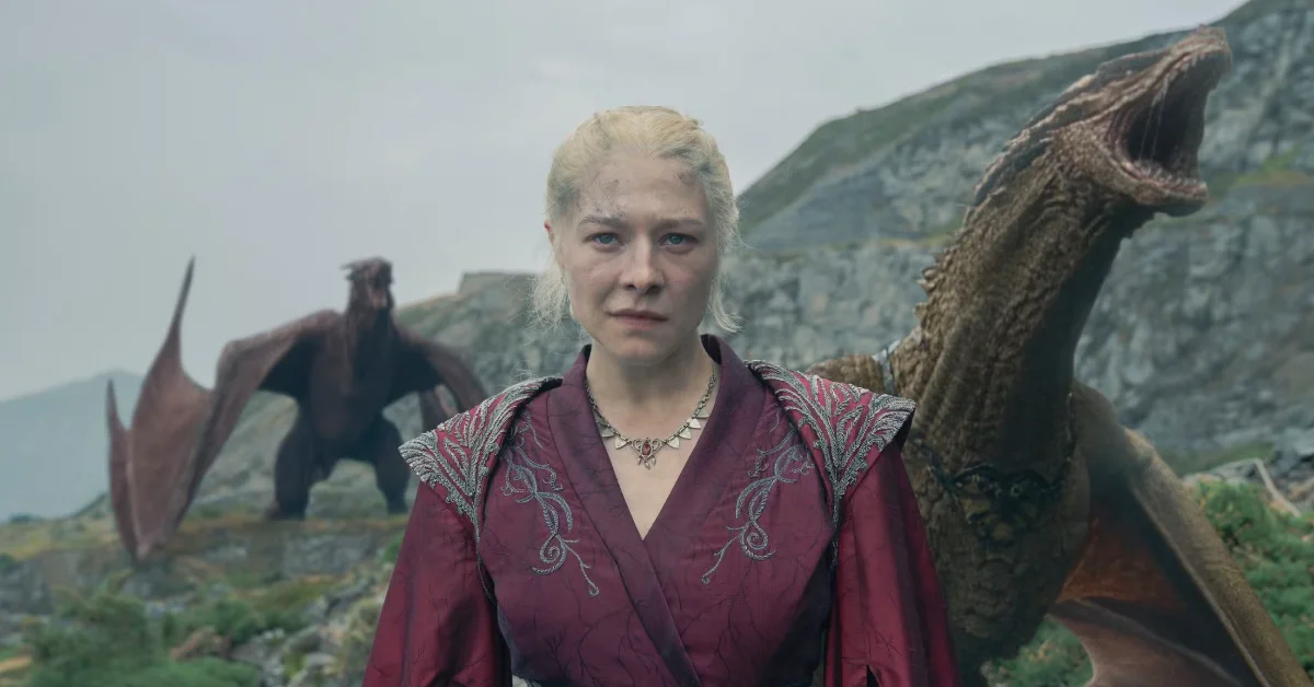 A still from House of the Dragon: Rhaenyra, a woman with platinum blonde hair wearing a burgundy tunic with embroidered shoulders and a metal necklace, stands in a mountainous landscape with large scaled reptilian creatures behind her.