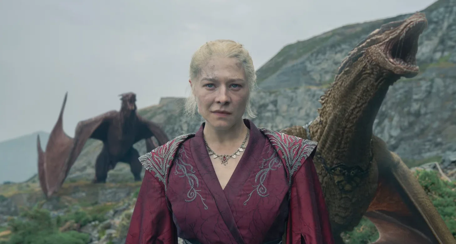 A still from House of the Dragon: Rhaenyra, a woman with platinum blonde hair wearing a burgundy tunic with embroidered shoulders and a metal necklace, stands in a mountainous landscape with large scaled reptilian creatures behind her.
