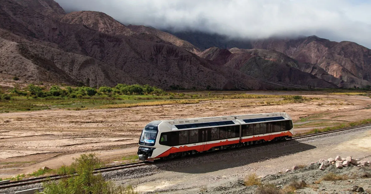 An orange and white train travels on tracks through a wide valley with colorful striped mountains in the background under cloudy skies.