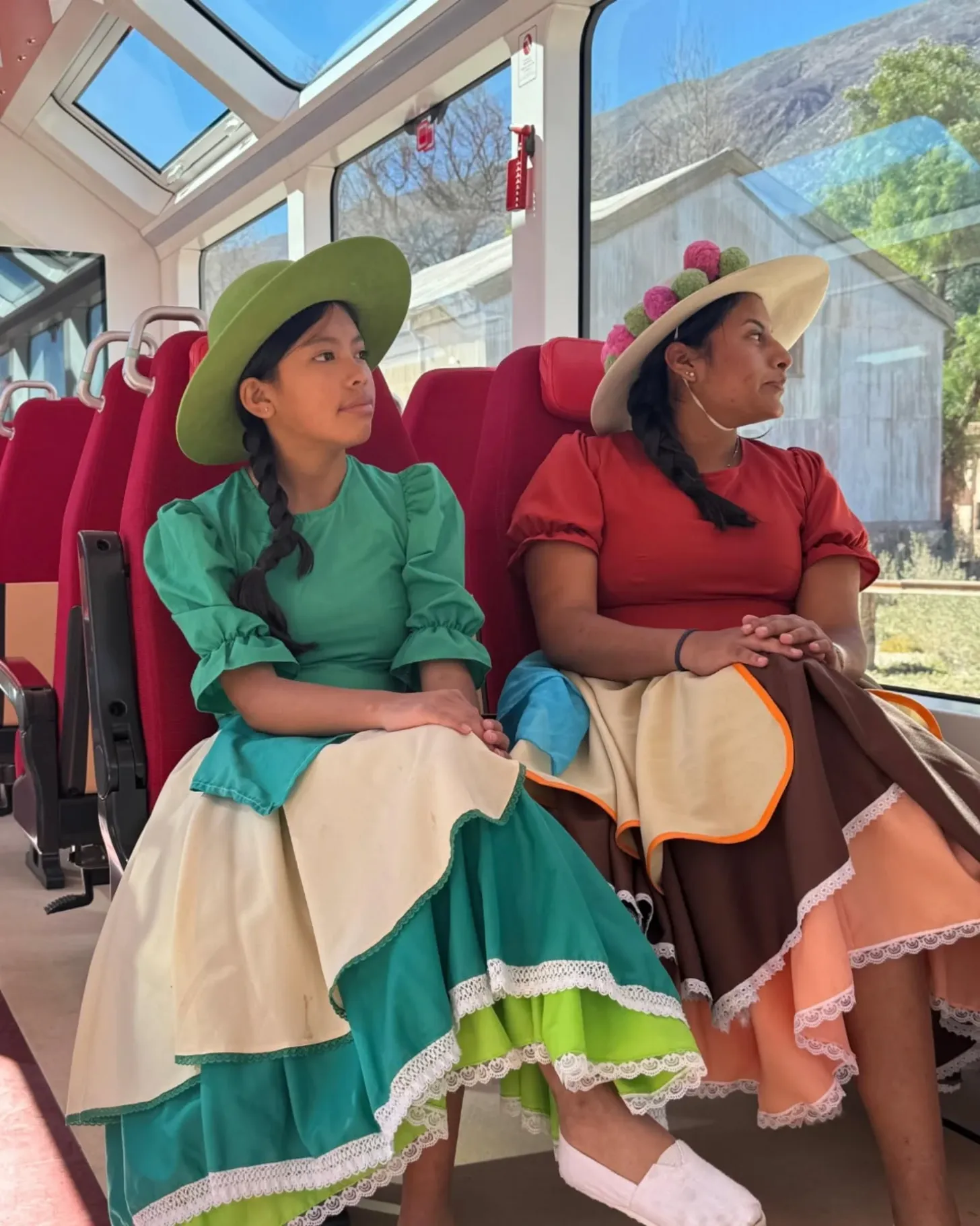Two women wearing wide-brimmed hats and colorful traditional dresses sit on red seats inside a train with large windows overlooking a mountainous landscape.