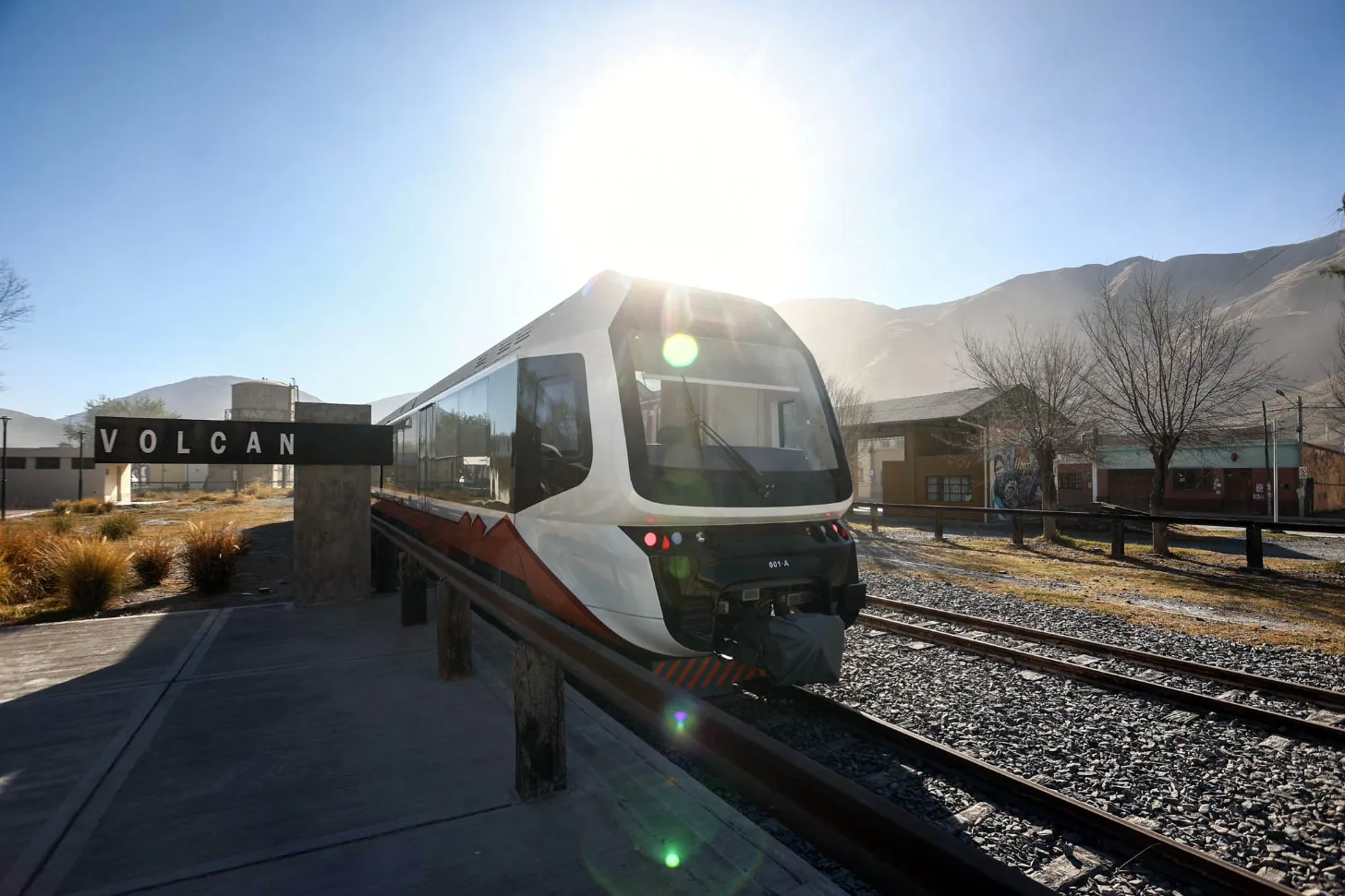 A modern white and orange train stopped at Volcan station, with mountains visible in the background under bright sunlight.