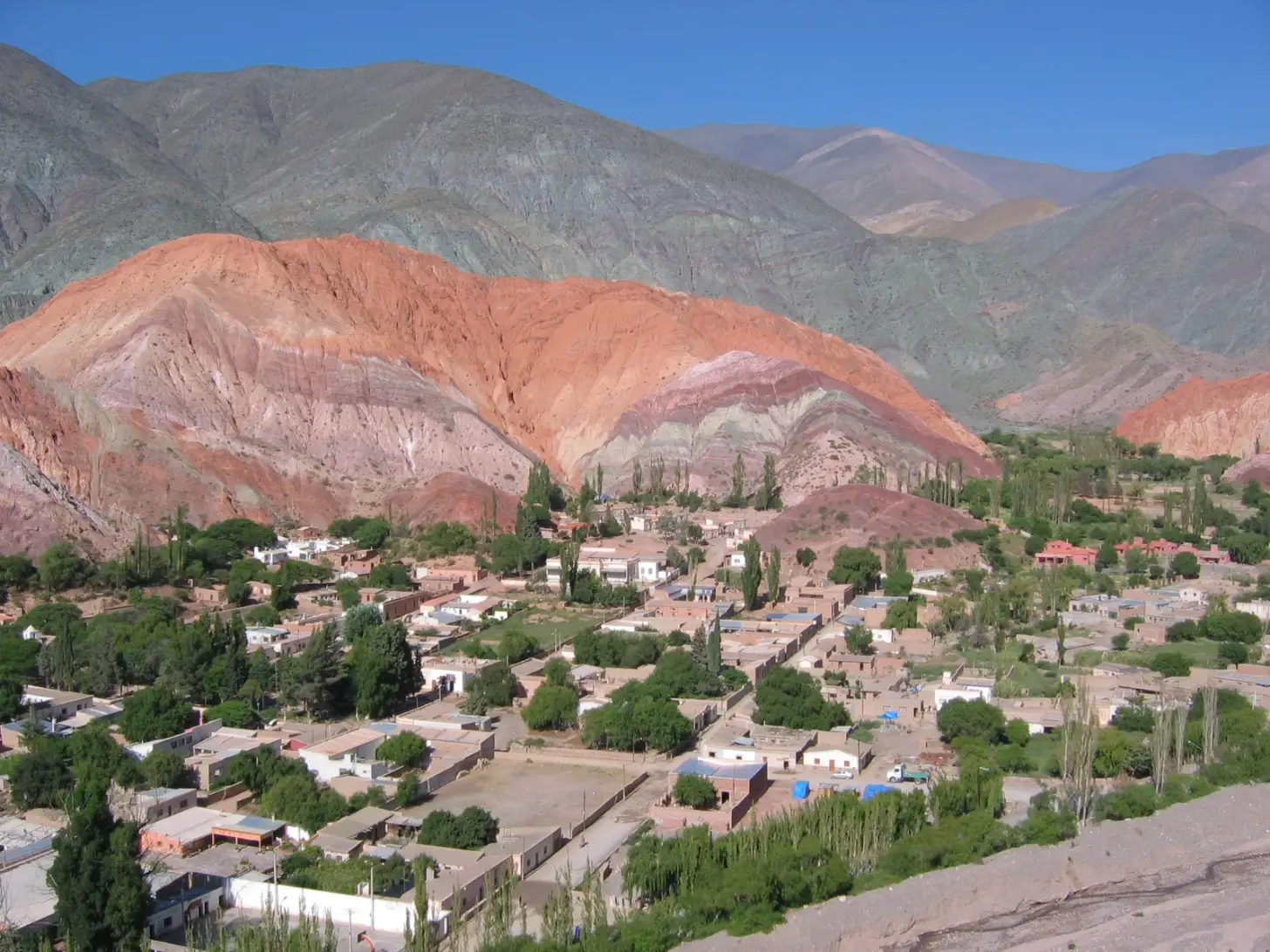 A town nestled in a valley surrounded by colorful striped mountains in shades of red, pink, and gray. White and tan buildings with flat roofs cluster among green trees, with tall cypress trees scattered throughout the settlement.