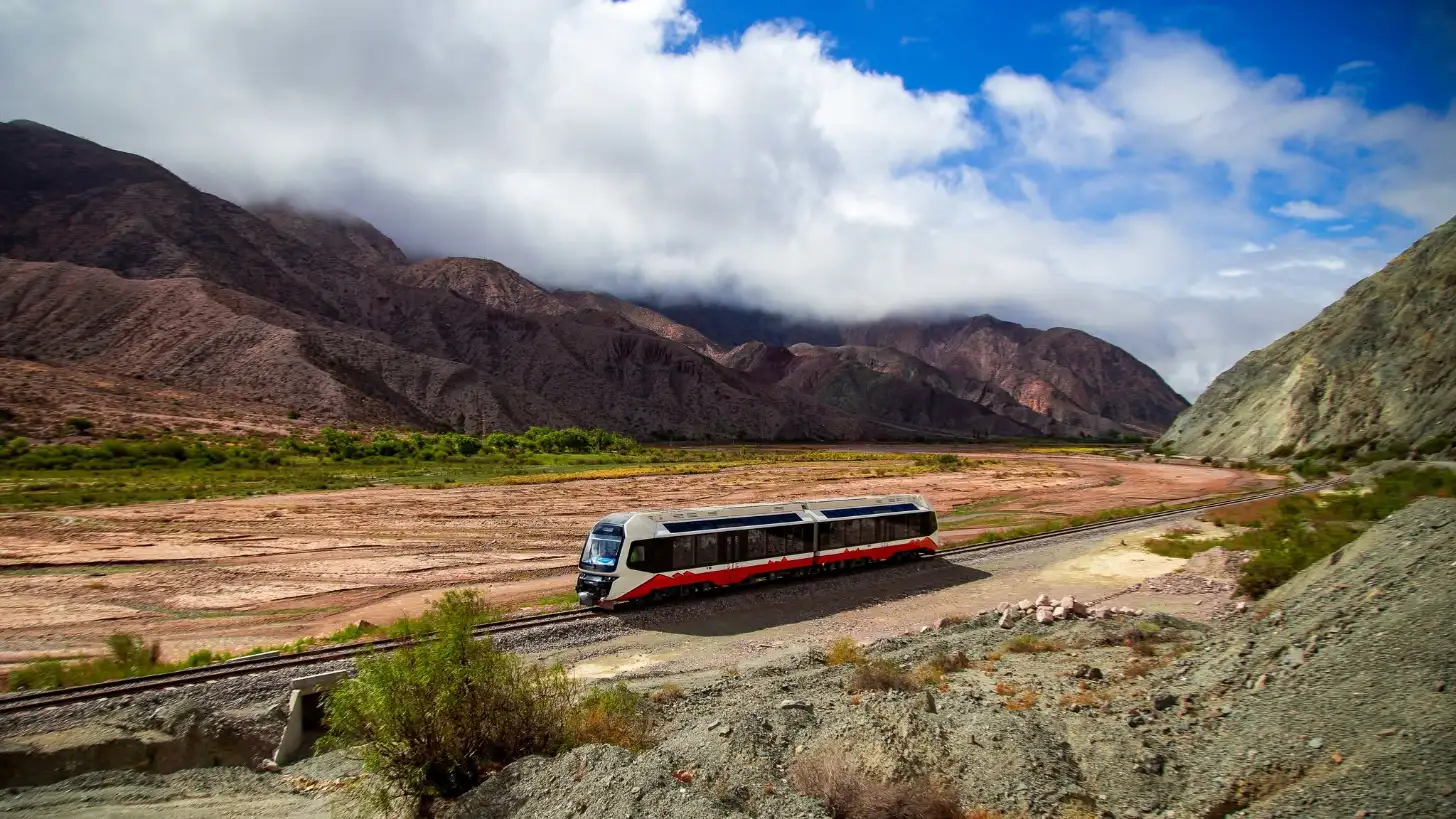 An orange and white passenger train travels on tracks through a valley with barren brown mountains on either side, green vegetation along the riverbank, and a blue sky with white clouds above.