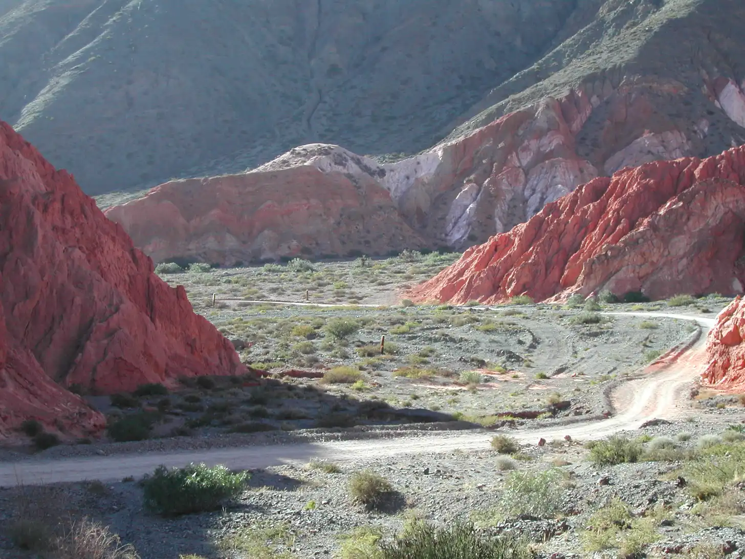 A road cuts through a valley floor dotted with sparse vegetation, flanked by steep red and white striped rock formations rising on both sides under a clear sky.