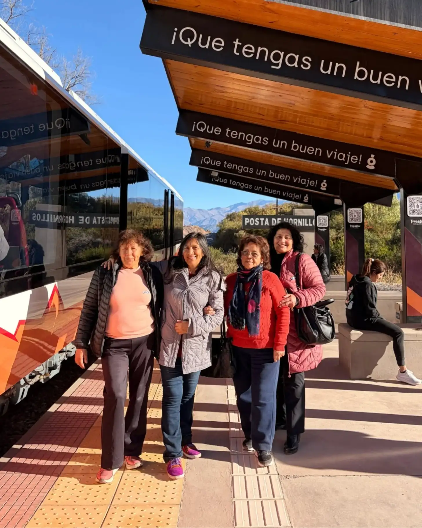 Four women stand together at a train station under a wooden shelter with Spanish text reading "¿Que tengas un buen viaje!" Mountains are visible in the background under clear blue sky.