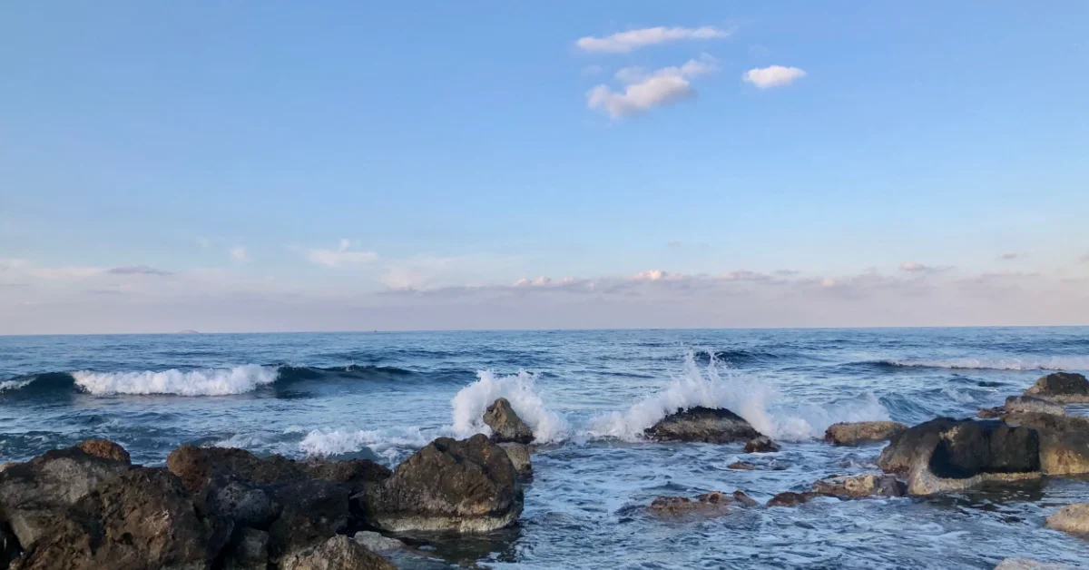 Ocean waves crash against dark rocks in shallow water under a blue sky with white clouds.