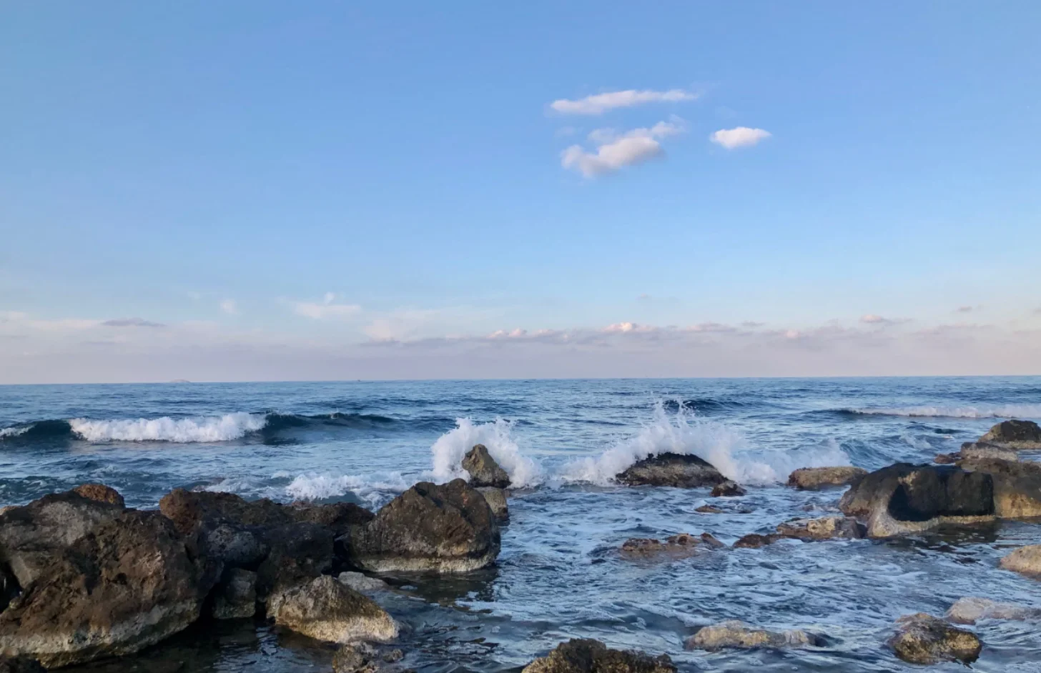 Waves crash against rocky outcrops along a coastline under a partly cloudy blue sky.