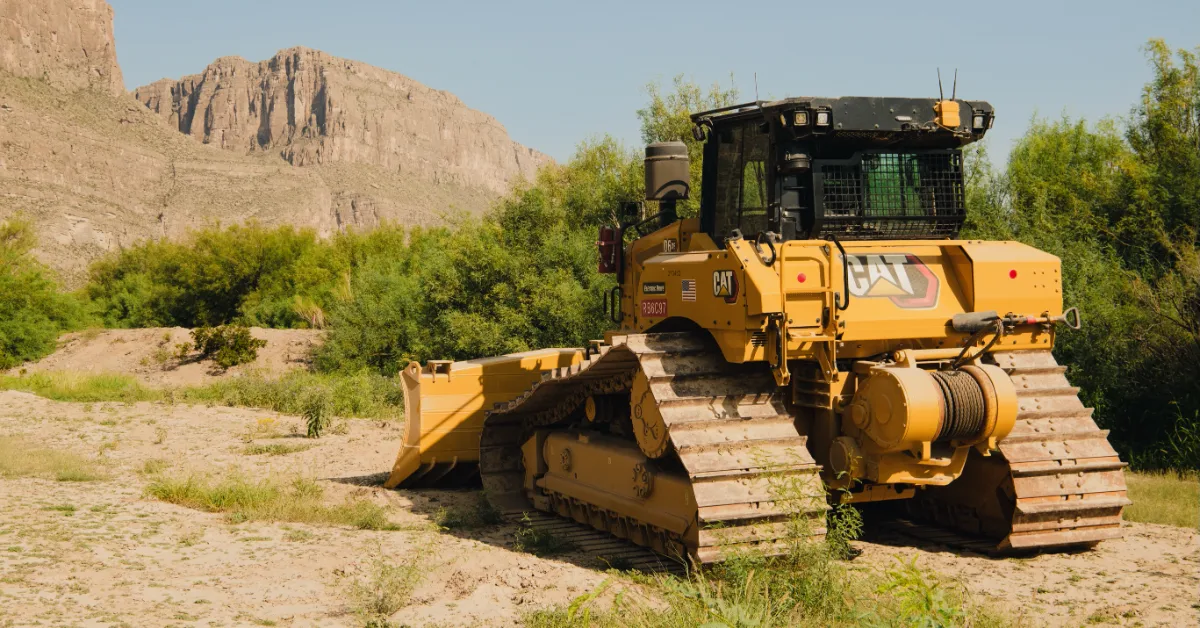 Yellow CAT bulldozer with tracked treads and front blade on sandy ground, rocky mountain visible in background, green vegetation surrounding the equipment.