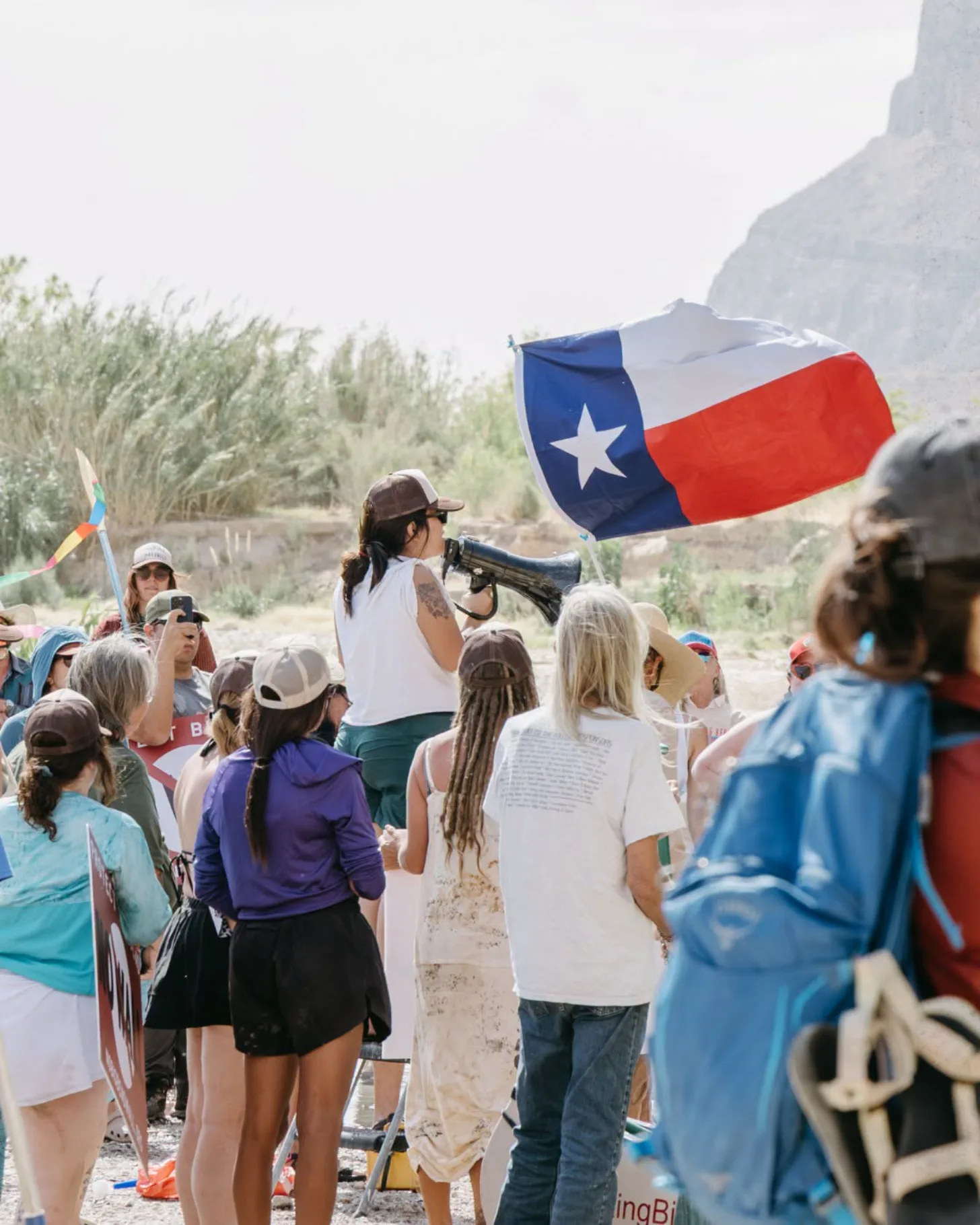A group of people gather outdoors near a rocky cliff, with one person on elevated ground holding a large Texas flag. Various other flags are visible among the crowd, and desert vegetation surrounds the scene.