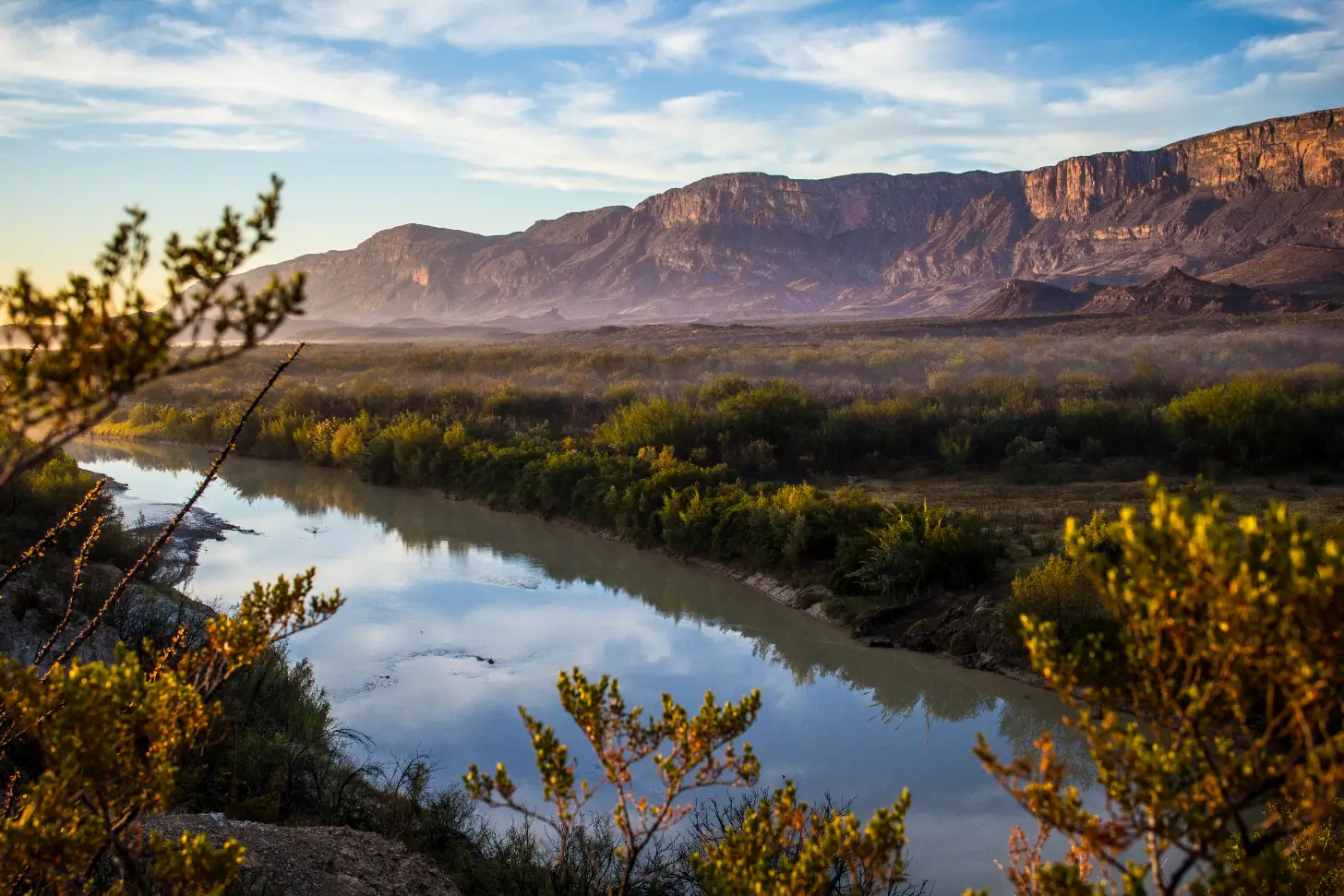 A river winds through desert scrubland with dense vegetation lining its banks, reflecting the blue sky. Tall, layered mountains with reddish-brown cliffs rise across the valley under partly cloudy skies.