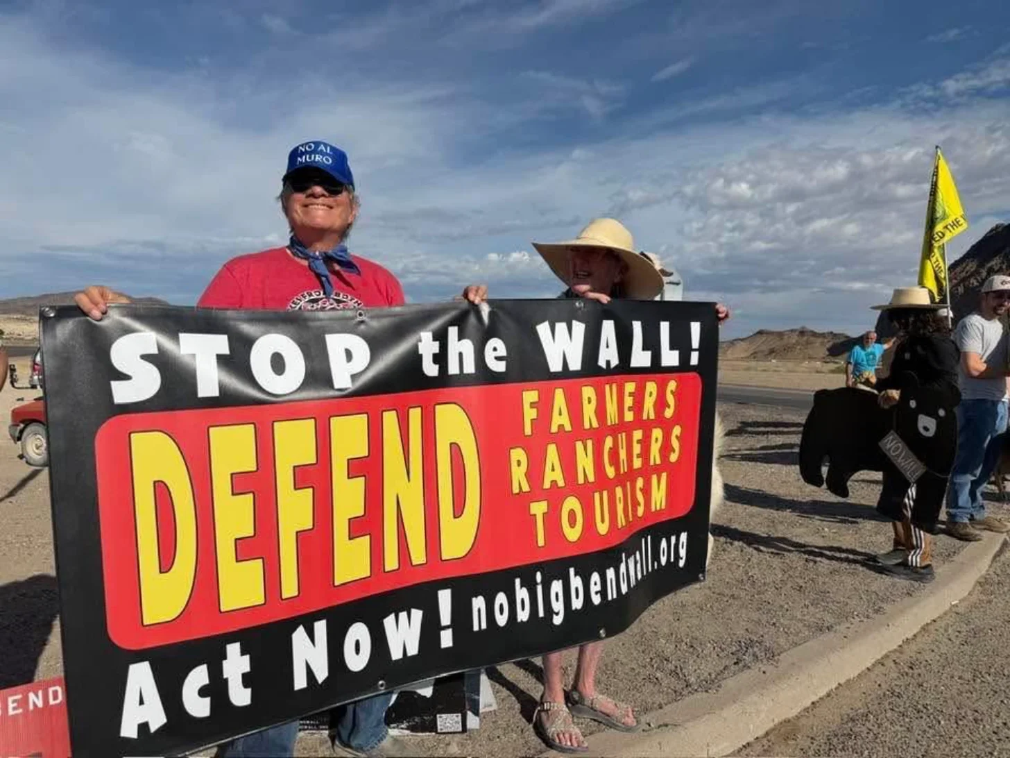 Two people hold a protest banner reading "STOP the WALL! DEFEND FARMERS RANCHERS TOURISM Act NOW! nobigbendwall.org" at an outdoor rally in an arid landscape.