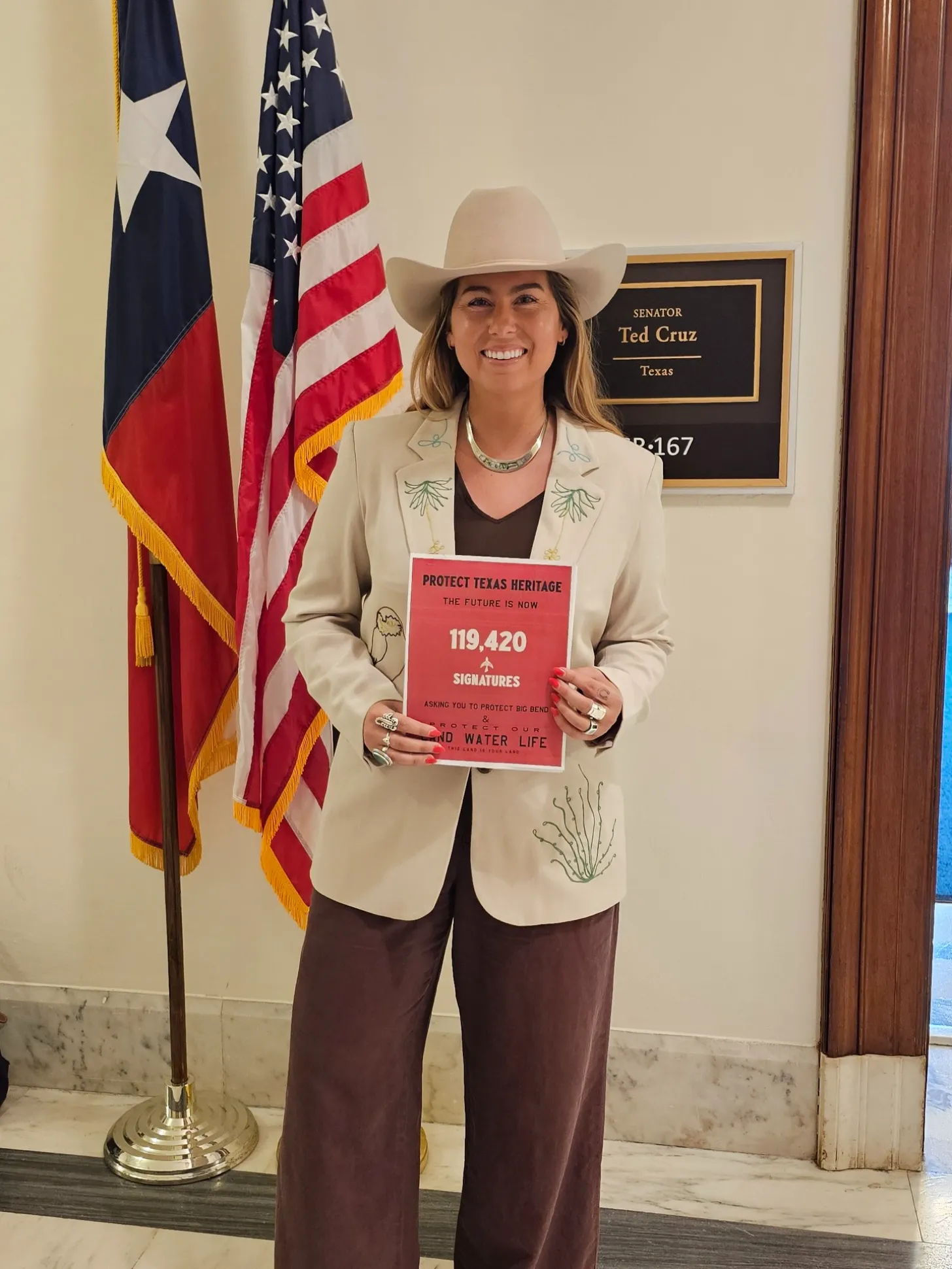 Woman in cream blazer and cowboy hat holds red sign reading "PROTECT TEXAS HERITAGE THE FUTURE IS NOW 119,420 SIGNATURES" in an office with Texas and U.S. flags and a nameplate for Senator Ted Cruz of Texas.