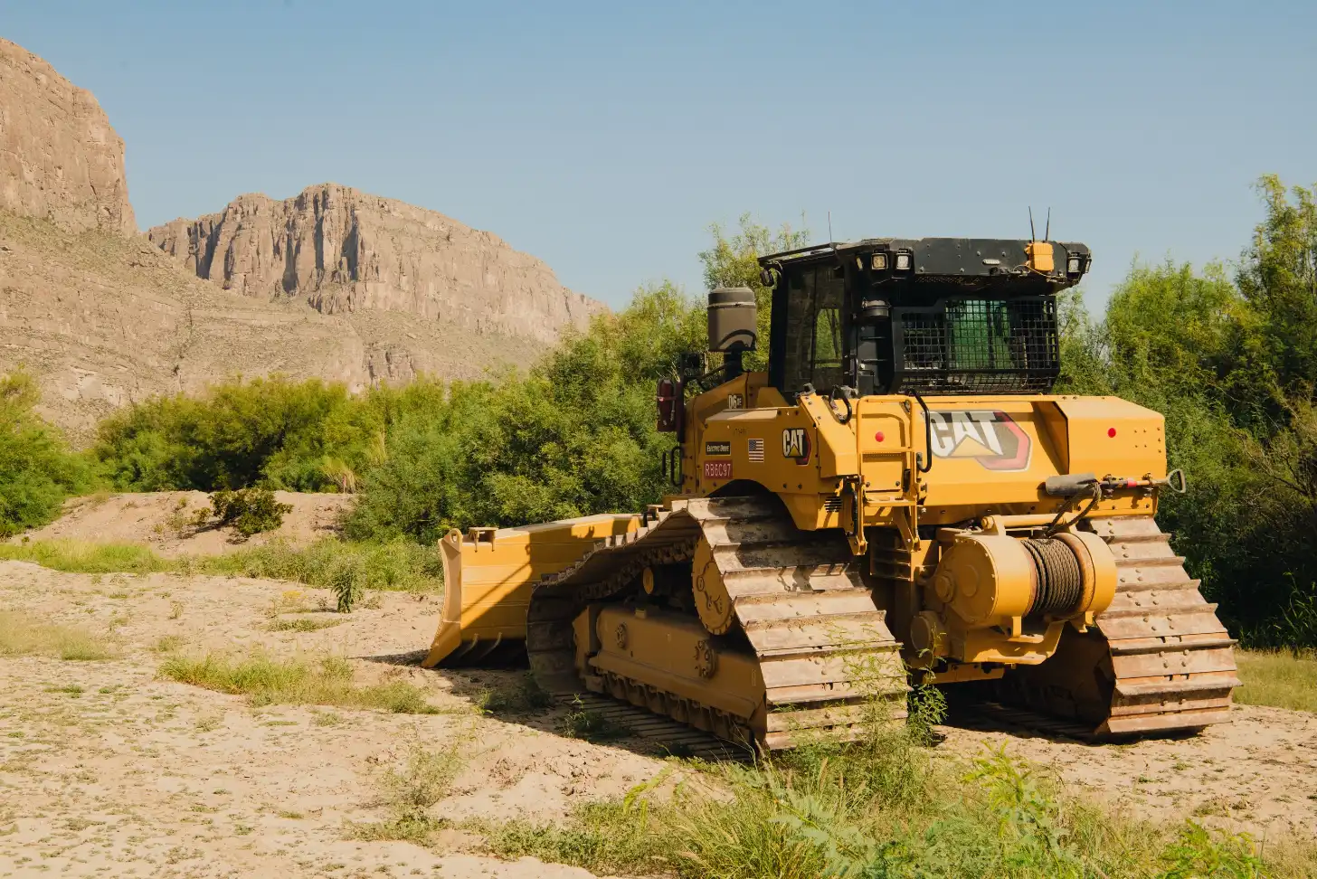 A yellow CAT bulldozer with tracked treads and a blade attachment sits on sandy ground with green shrubs and a rocky cliff formation in the background.