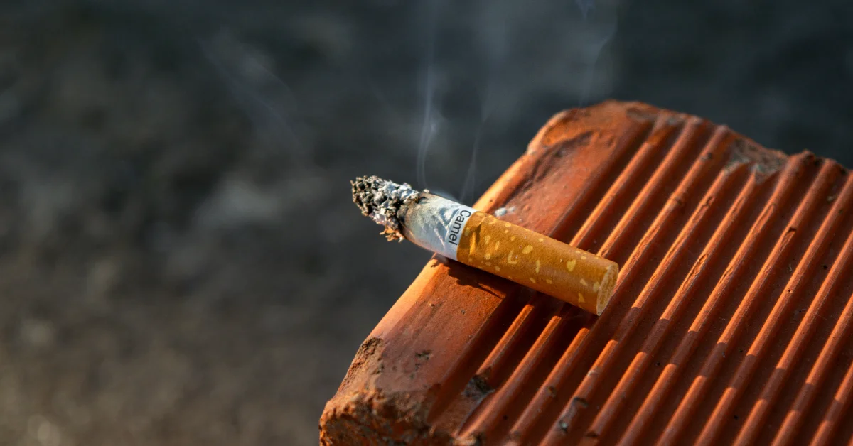 A lit cigarette rests on stacked terra cotta roof tiles against a dark blurred background, with smoke rising from its tip.