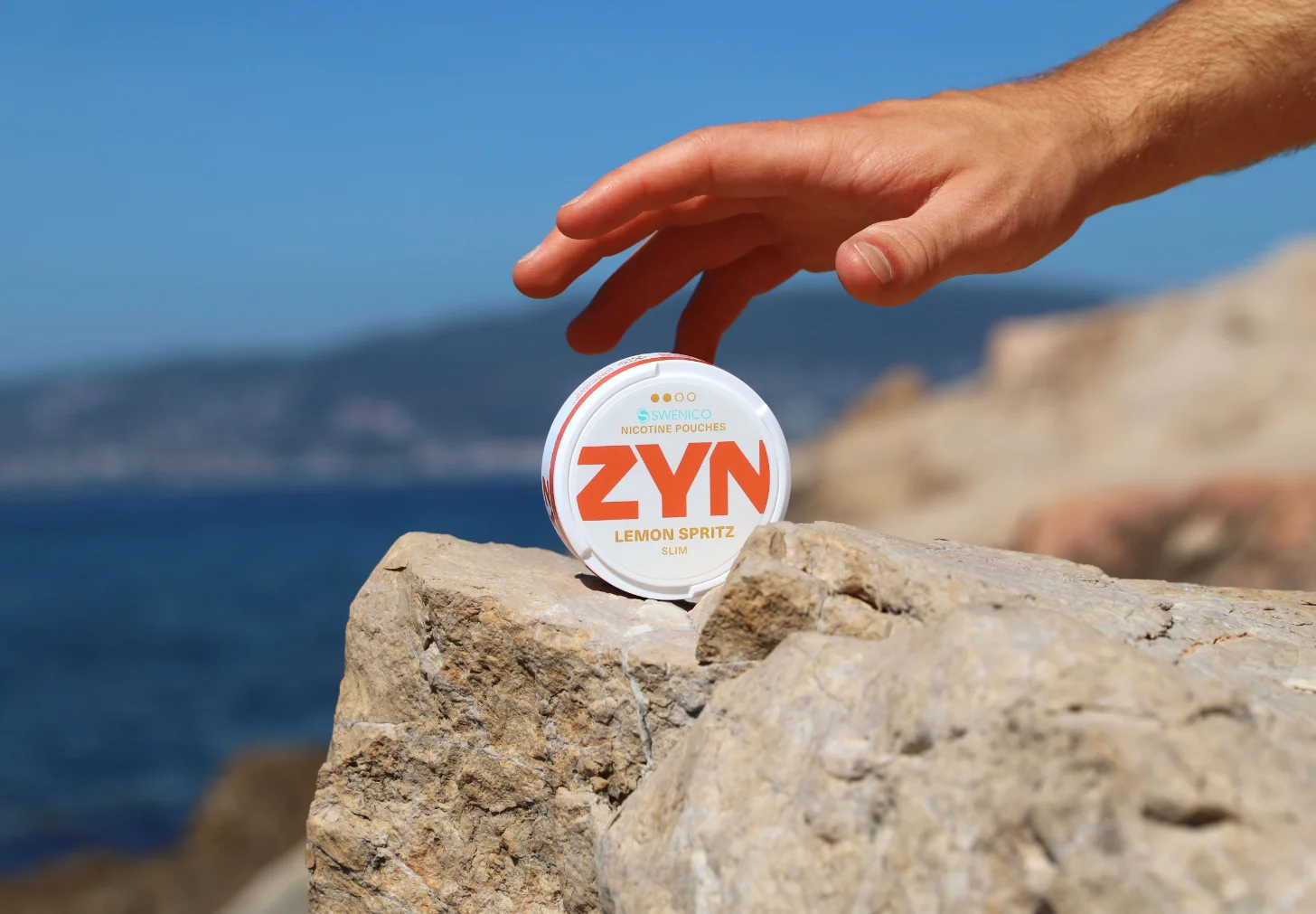 A hand reaches toward a white circular nicotine pouch container labeled "ZYN LEMON SPRITZ SLIM" positioned on a rocky surface, with a blue ocean and coastline visible in the blurred background.