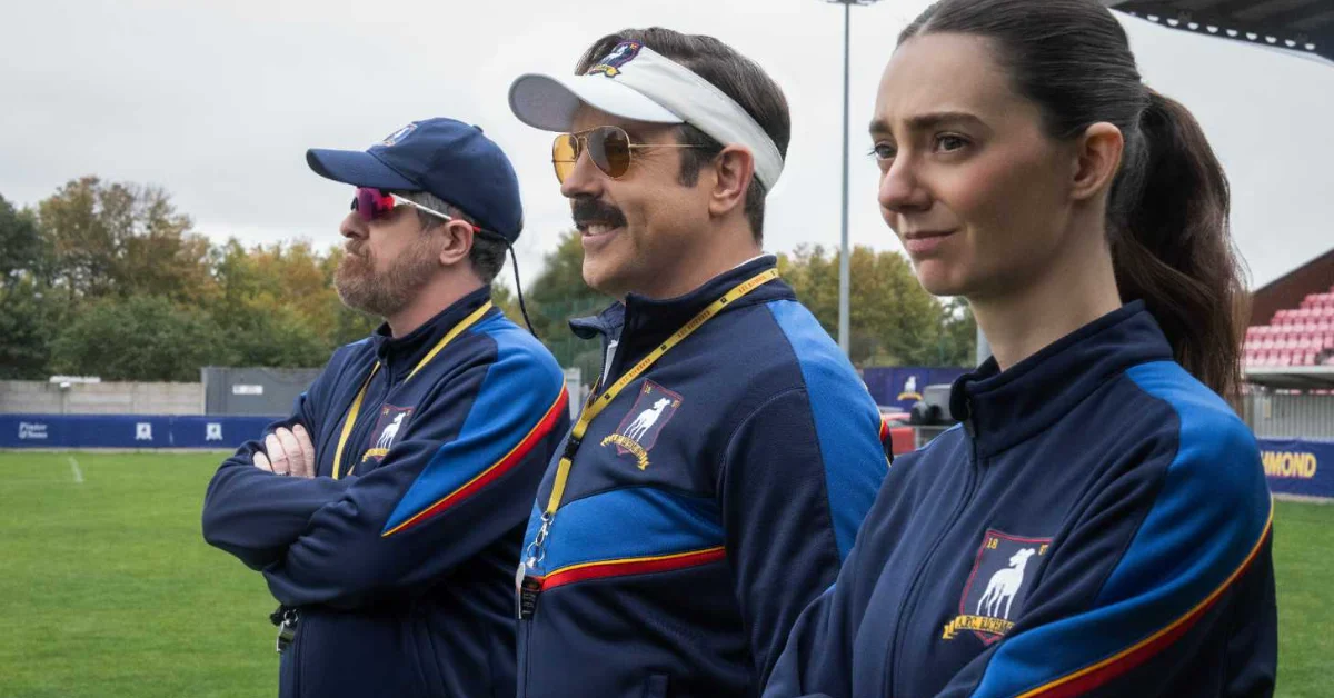 A still from "Ted Lasso:" Three people in matching navy and blue jackets with yellow and red stripes stand on a sports field. The person in the center wears a white visor and gold-tinted sunglasses and has a medal around their neck. Trees and stadium seating are visible in the background.
