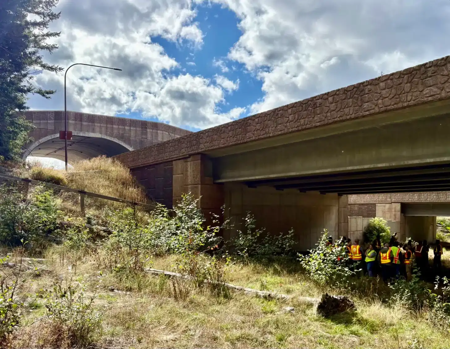 Concrete overpass with brown stone facade spanning overgrown vegetation and sandy ground; group of people in safety vests visible beneath the structure against blue sky with white clouds.