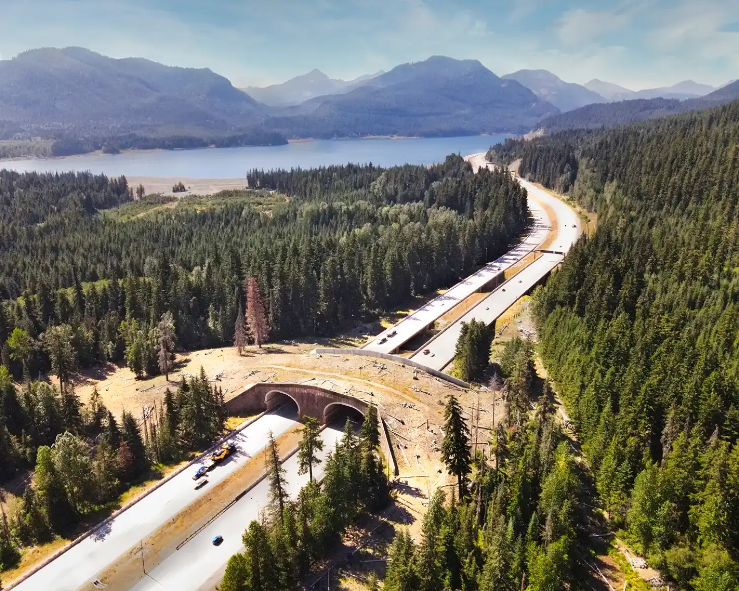 Aerial view of a highway with two parallel white lanes running through dense coniferous forest, with concrete tunnels visible beneath the road surface. A large blue lake surrounded by forested mountains appears in the background under a partly cloudy sky.