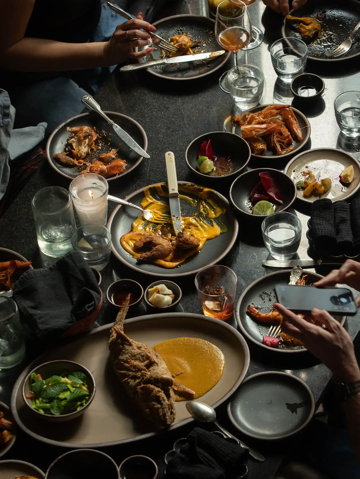 A dark table set with multiple plates of cooked meat dishes, roasted vegetables, and small bowls of condiments. Several diners' hands are visible holding utensils and a phone while eating. Glasses of water and candles are placed around the table.