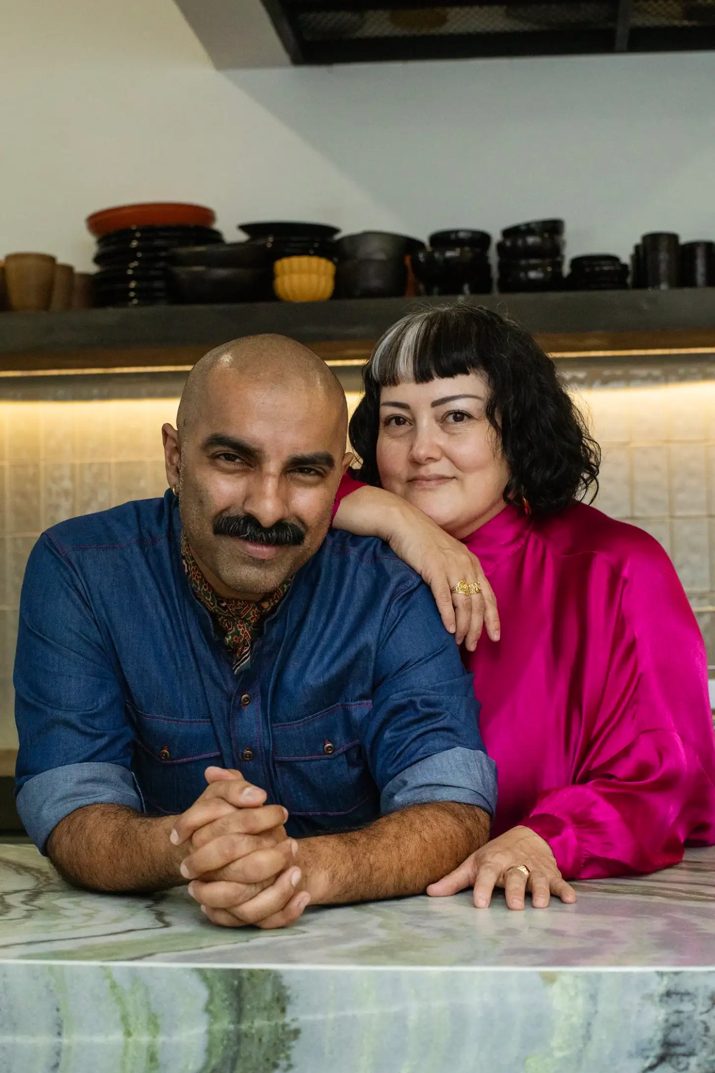 Two people lean against a counter in what appears to be a kitchen or studio space. The man on the left wears a blue denim shirt and has a full beard. The woman on the right wears a bright pink shirt and has short black hair with bangs. Behind them, shelves display various black ceramic vessels and containers.