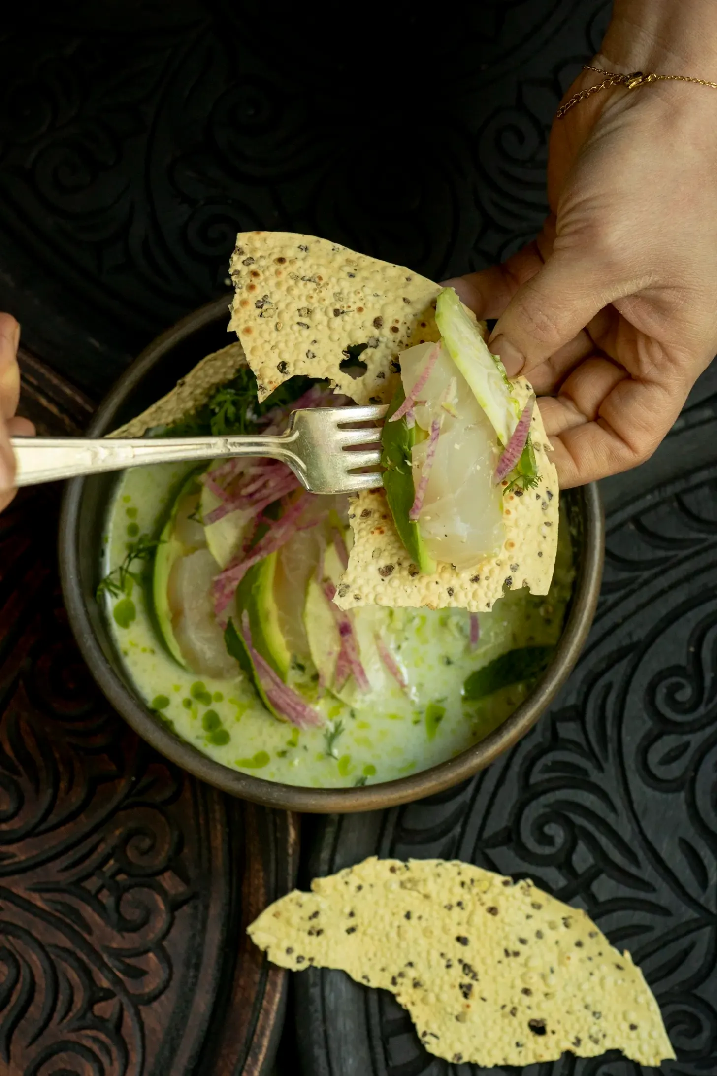 A hand holds a seeded tortilla with a fork over a bowl containing a creamy pale green soup or dip garnished with microgreens and pickled onions. Additional seeded tortillas rest on an ornately patterned dark surface beside the bowl.