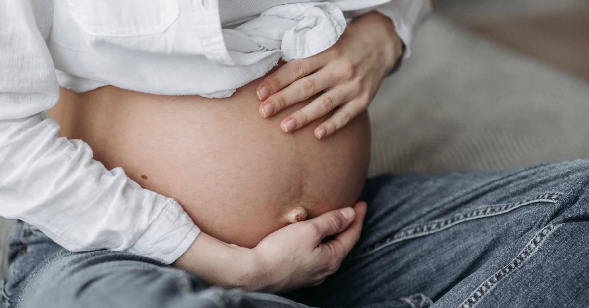 A pregnant person wearing a white shirt and jeans holds their bare belly with both hands.