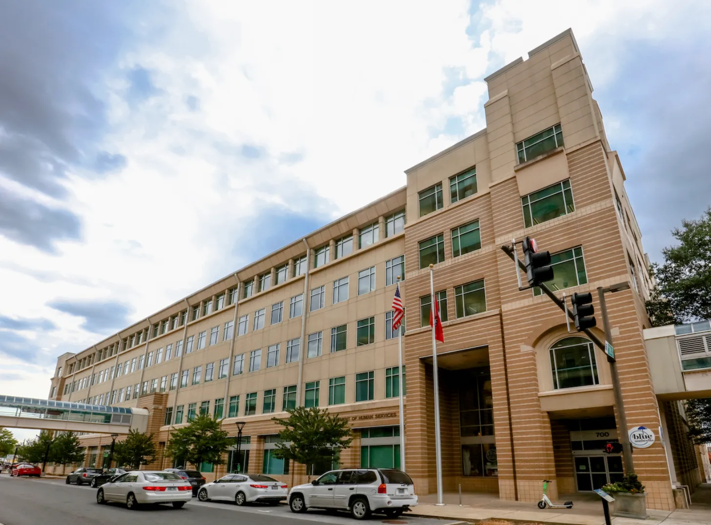 Brick and glass mid-rise building with multiple floors and windows, American flag and red flag hanging near entrance, parked cars in front, traffic light visible, trees and cloudy sky.