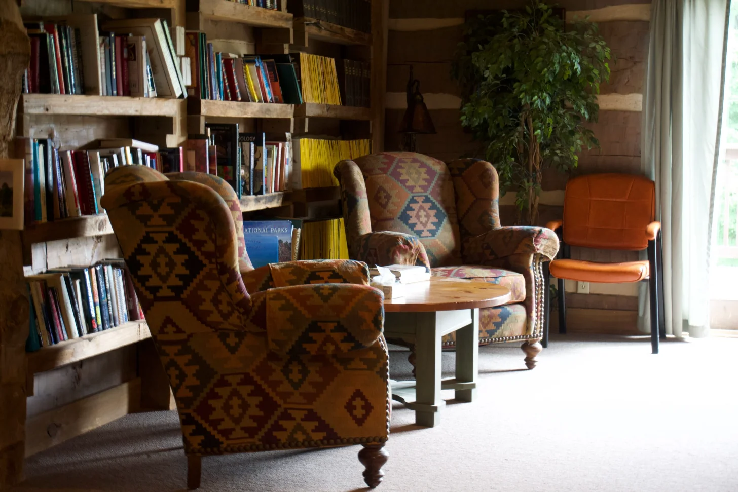 A library lounge with patterned upholstered armchairs arranged around a wooden table, wooden bookshelves filled with books along the walls, a potted plant, and an orange chair visible on the right side.