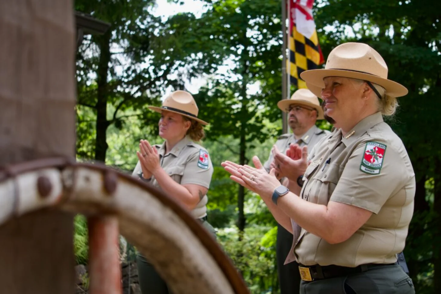 Three park rangers in tan uniforms and campaign hats stand outdoors clapping, with a Maryland flag visible in the background and trees behind them.