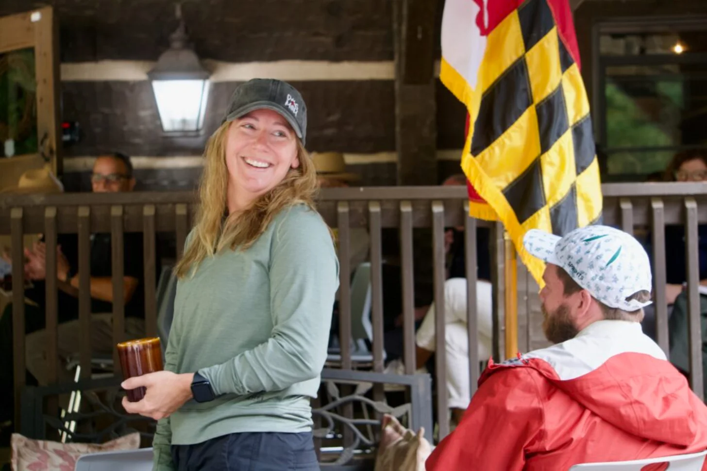 Woman in gray long-sleeve shirt and dark cap holding a beverage smiles at the camera while seated next to a man in a red jacket and white cap, with a Maryland state flag visible in the background.