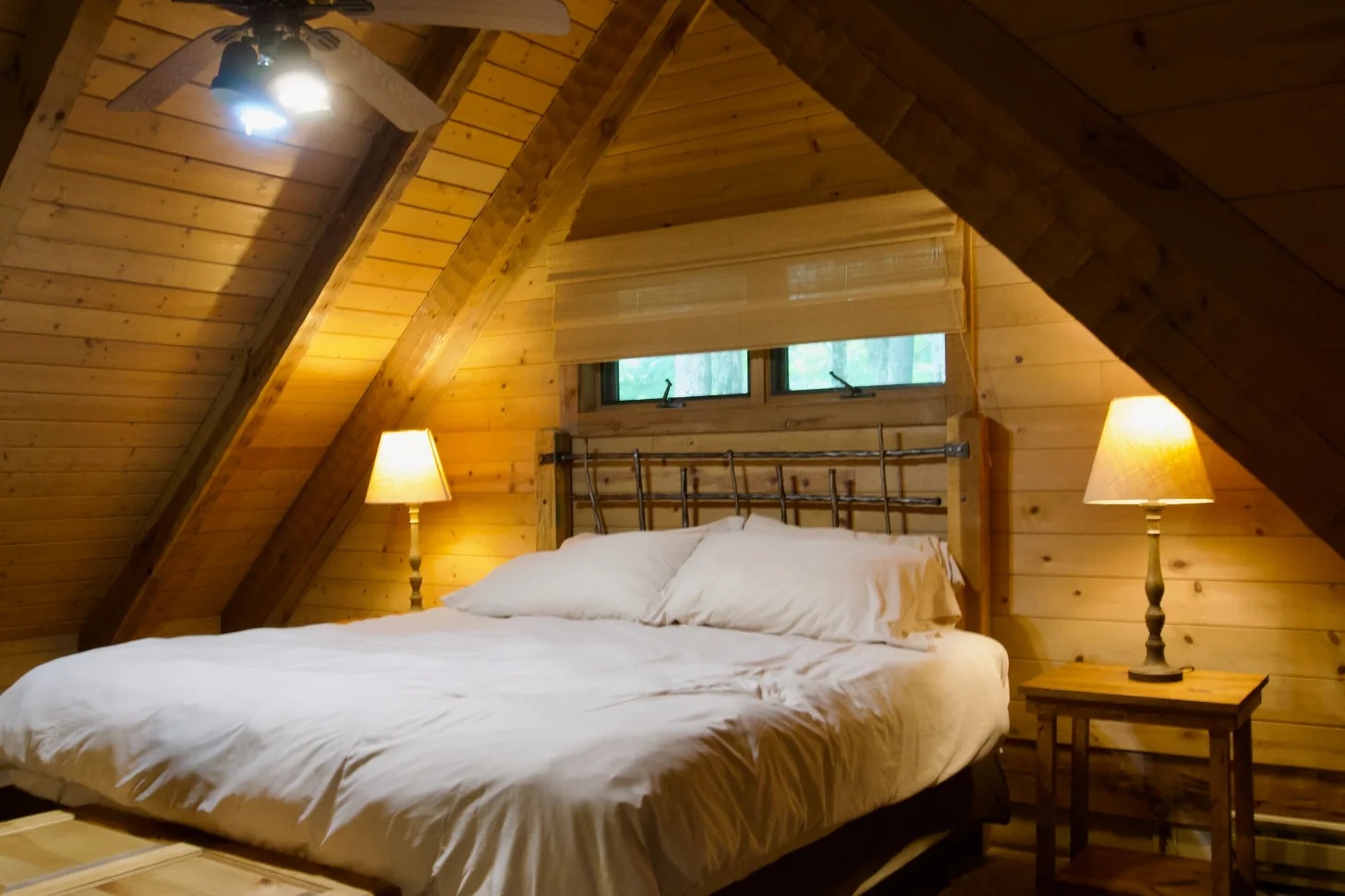 A bed with white pillows and bedding sits in an A-frame wooden cabin bedroom with knotty pine walls and ceiling. Two table lamps with cream shades flank the bed, and small windows above the headboard look out to trees. A ceiling fan with light fixture is mounted overhead.
