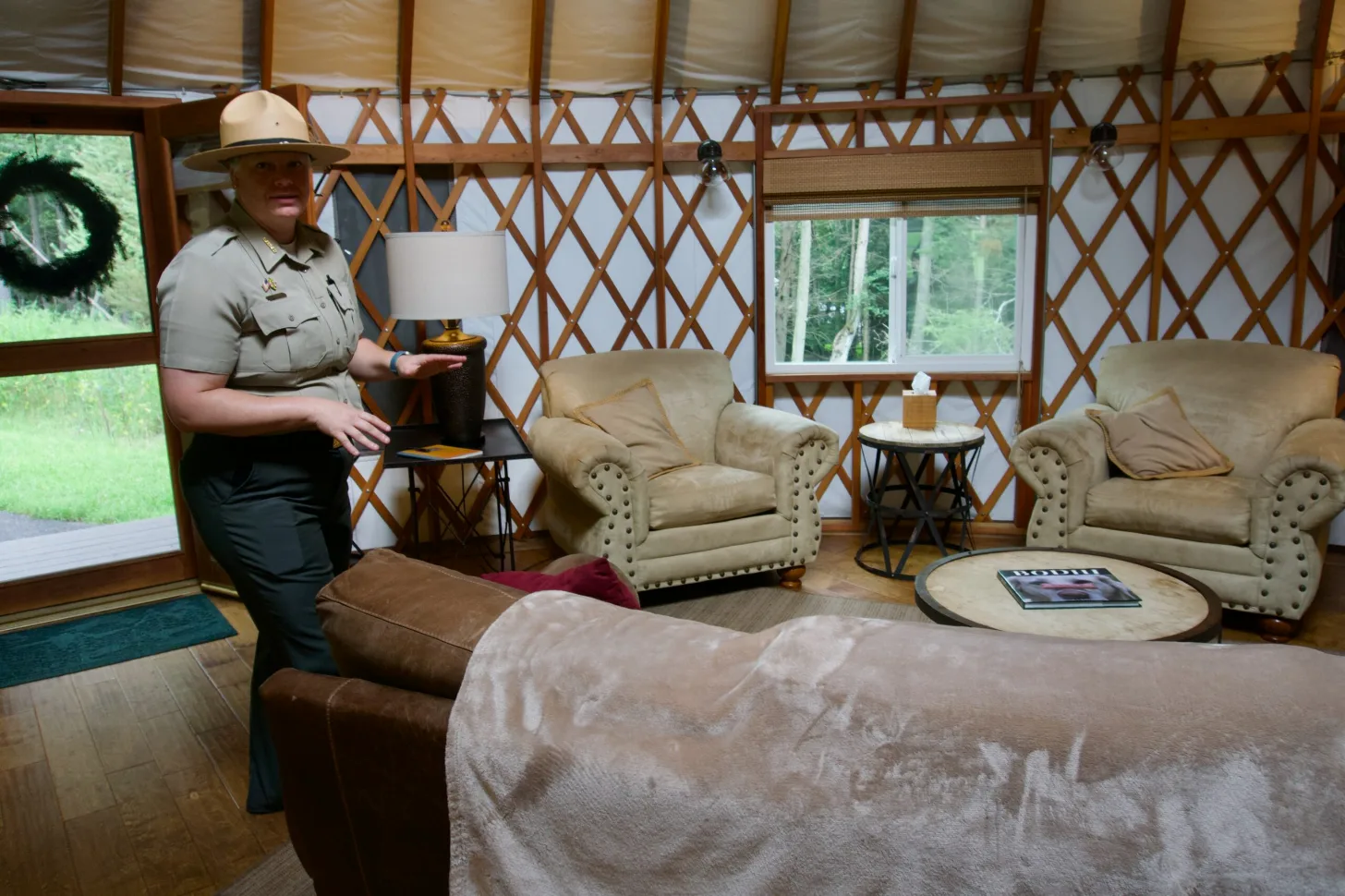 Park ranger in uniform stands in a yurt interior with beige armchairs, a brown sofa, wooden lattice walls, and windows overlooking forest.