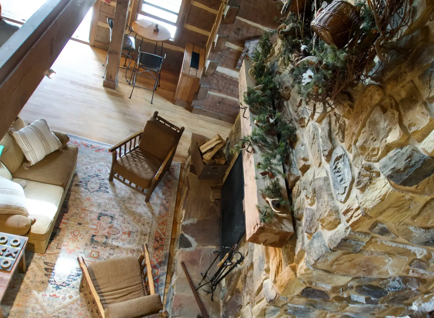 Overhead view of a multi-level interior space with wooden beams, stone walls covered in climbing ivy, wooden chairs and furniture on a patterned rug, and large windows allowing natural light.