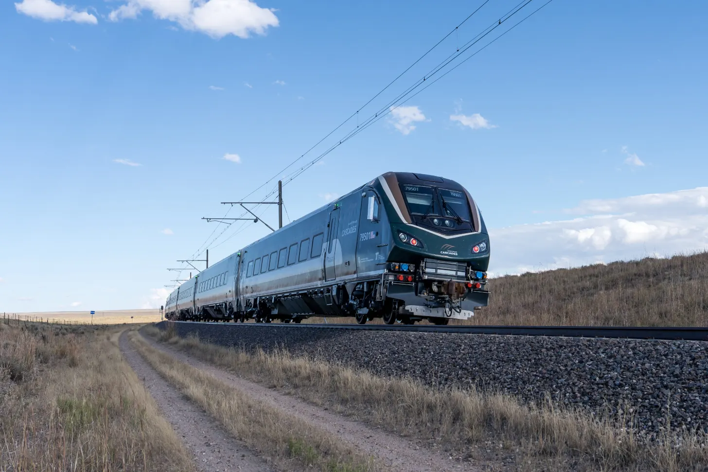 A silver and dark green passenger train travels on railroad tracks through an arid landscape with sparse brown vegetation and distant hills under a partly cloudy sky.