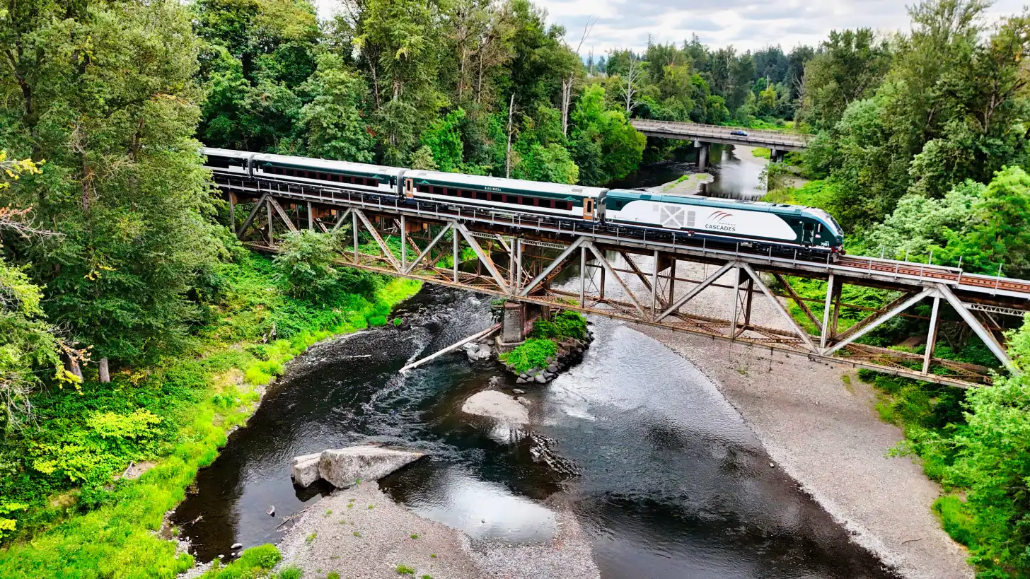 A passenger train labeled "Cascades" crosses a wooden trestle bridge over a river surrounded by dense green forest and evergreen trees.