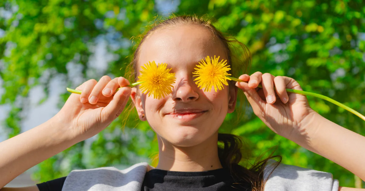 A child holds two yellow dandelions up to their eyes like glasses, smiling at the camera with green foliage blurred in the background.