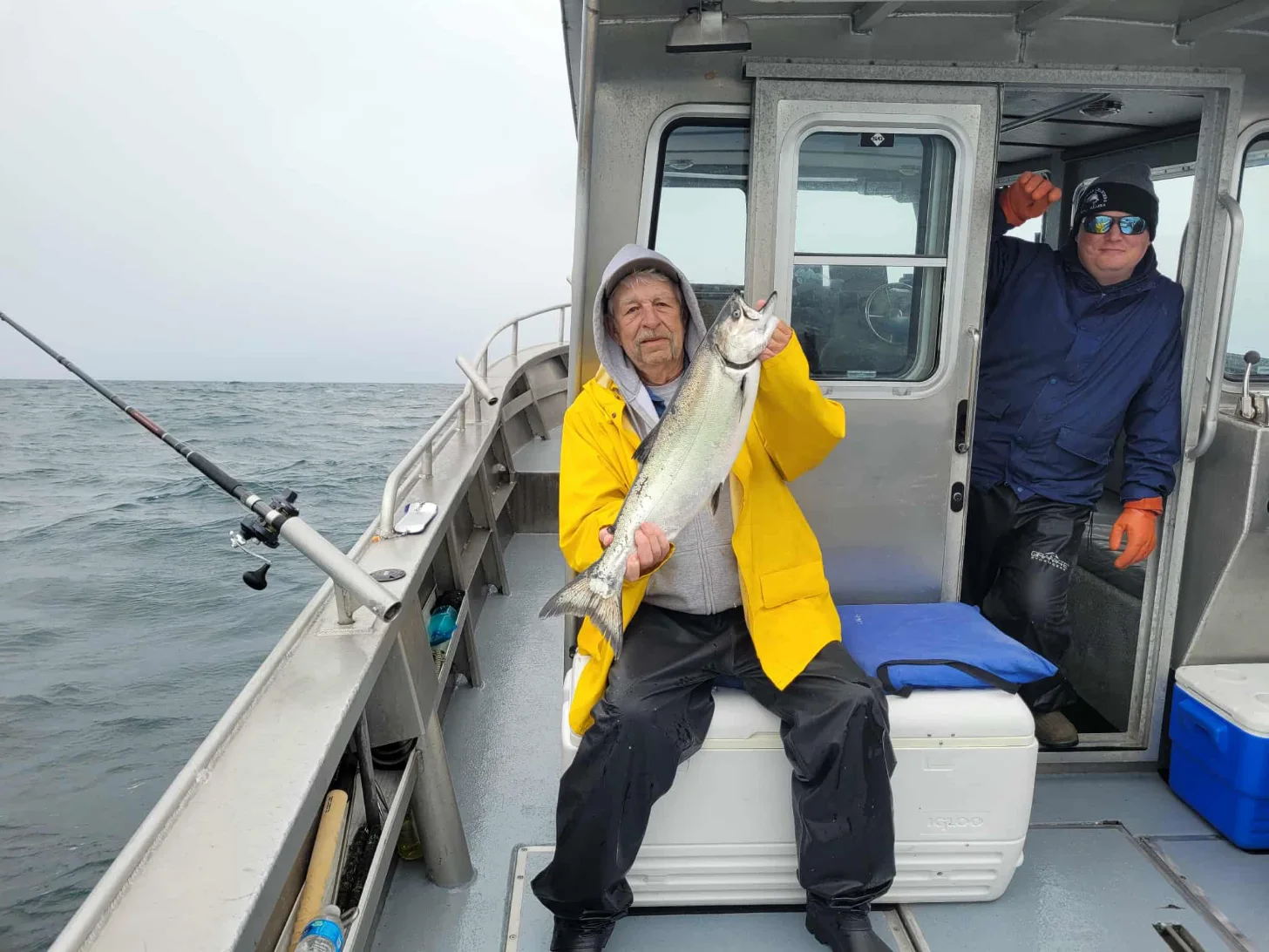 A man in a yellow jacket and white hood sits on a fishing boat holding a large silvery fish. Another man in dark clothing and sunglasses stands in the cabin doorway behind him.