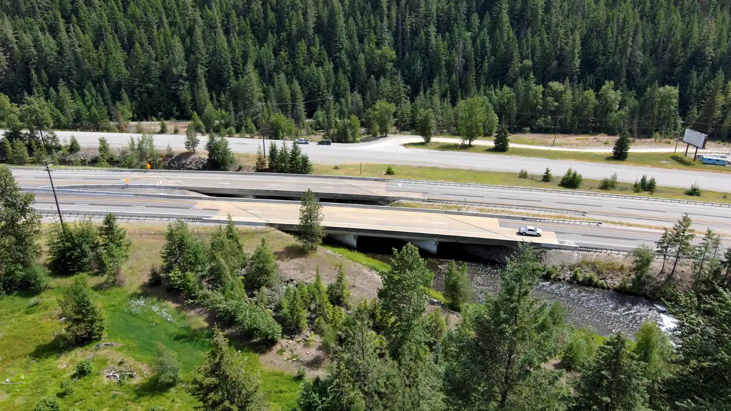 Highway overpass crossing a river with coniferous forest on both sides; a white vehicle travels on the road above.