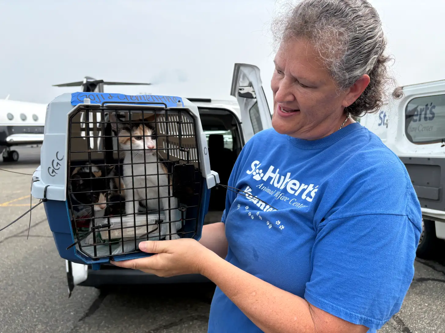 A woman wearing a blue St. Hubert's Animal Care Center shirt holds a transport carrier containing a cat, standing in front of a white van at an airport with a helicopter visible in the background.