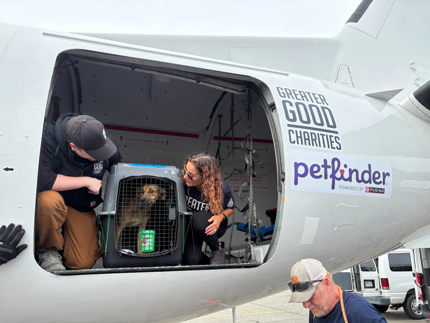 Two people load a pet carrier containing a brown dog into an aircraft cargo hold. The aircraft exterior displays "GREATER GOOD CHARITIES" and "petfinder POWERED BY PURINA" text. A man in a cap stands nearby on the tarmac.