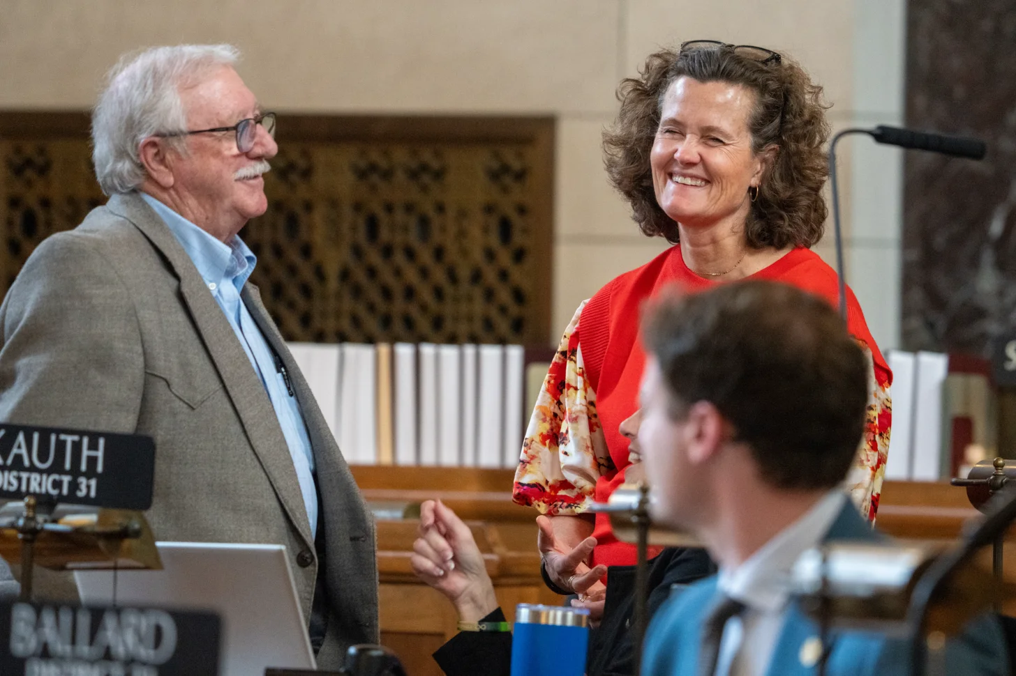 An older man in a gray blazer and blue shirt stands beside a woman in a red sweater with a floral scarf, both smiling during what appears to be a public meeting or event. A nameplate reading "AUTH DISTRICT 51" is visible on a table in the foreground.