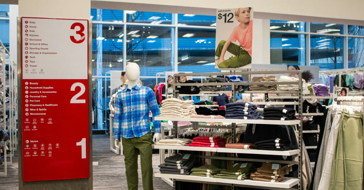 Inside a retail store, a mannequin wears a blue plaid shirt and olive pants while standing near display tables with folded clothing. A large red directory sign on the left lists store sections numbered 1, 2, and 3. Large windows show a parking structure outside.